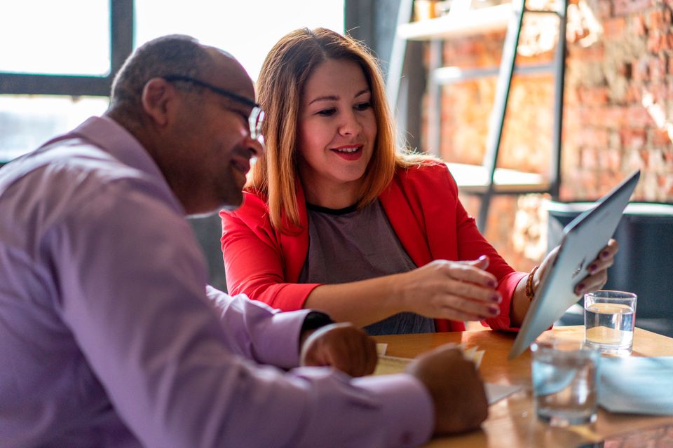 A woman in a red blazer walks a colleague through something on a tablet.