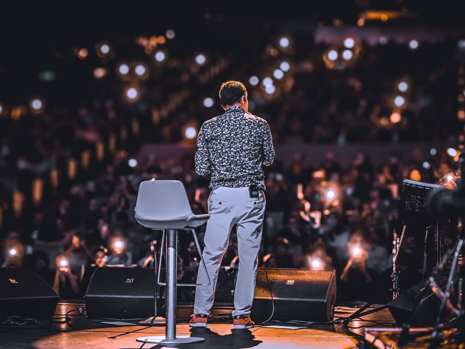 A speaker seen from behind on a lit stage, facing a full room.