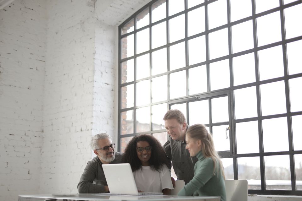 A small group gathers around a laptop beside a large window.