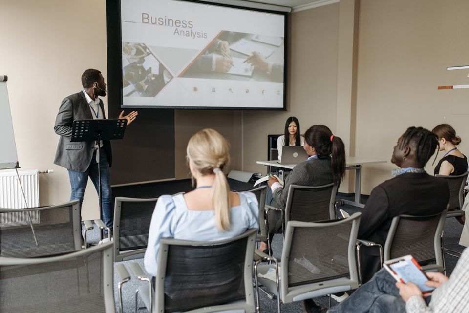 A presenter at a lectern speaks beside a projected slide to seated attendees.