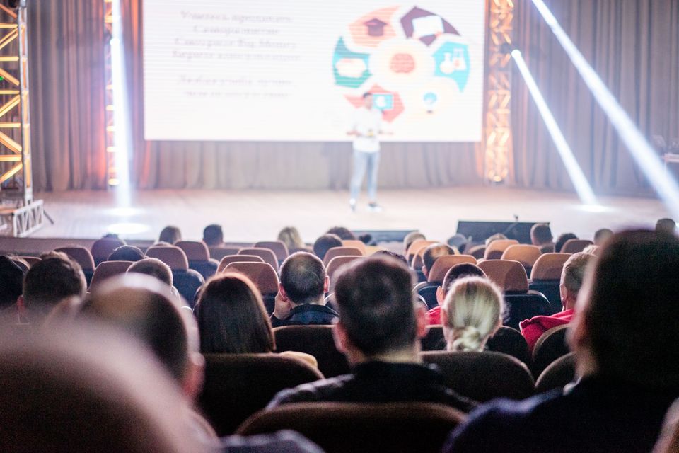 An audience seen from behind watches a presentation on a lit stage.